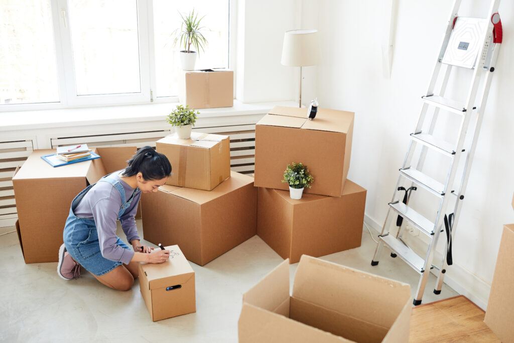 Wide angle background of woman packing cardboard boxes in empty white room, moving, relocation and house decor concept, copy space