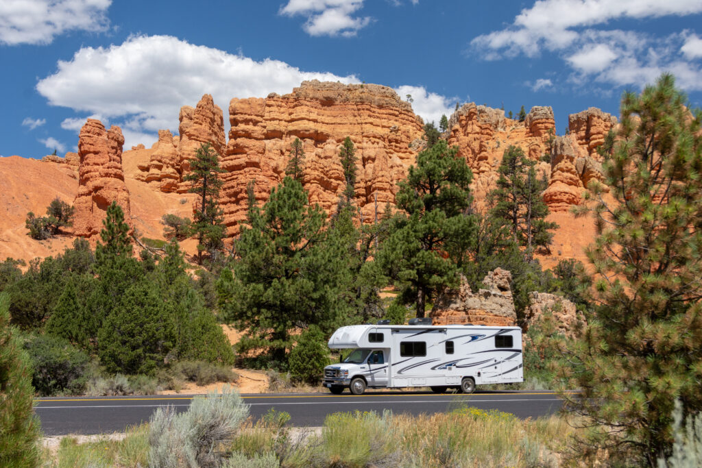 RV on road with green trees and red rock in the background.